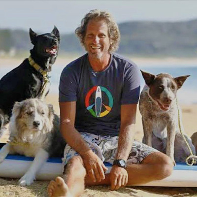 Chris sitting on the beach with his surf board and dogs.